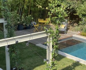 Jardin moderne avec piscine, terrasse en bois et mobilier de jardin jaune. Pergola végétalisée avec guirlande lumineuse devant un mur de bambous.