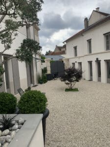 Cour moderne en gravier clair, bâtiments blancs aux volets gris. Un érable japonais, des oliviers, jardin paysager, portail foncé, vélo.