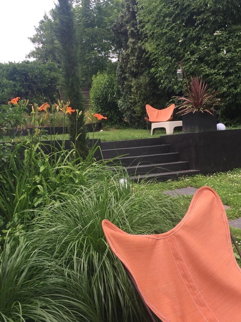 Jardin luxuriant avec chaises papillon oranges, hémérocalles éclatantes, grandes herbes vertes et escaliers sombres. Cadre naturel apaisant.