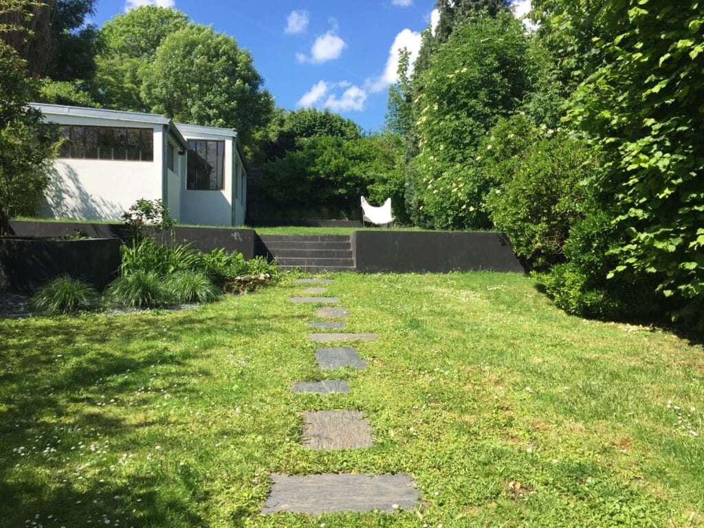 Jardin moderne et verdoyant avec un chemin de dalles, un escalier noir menant à une chaise papillon blanche, et un bâtiment épuré à gauche. Ciel bleu et arbres luxuriants en arrière-plan.
