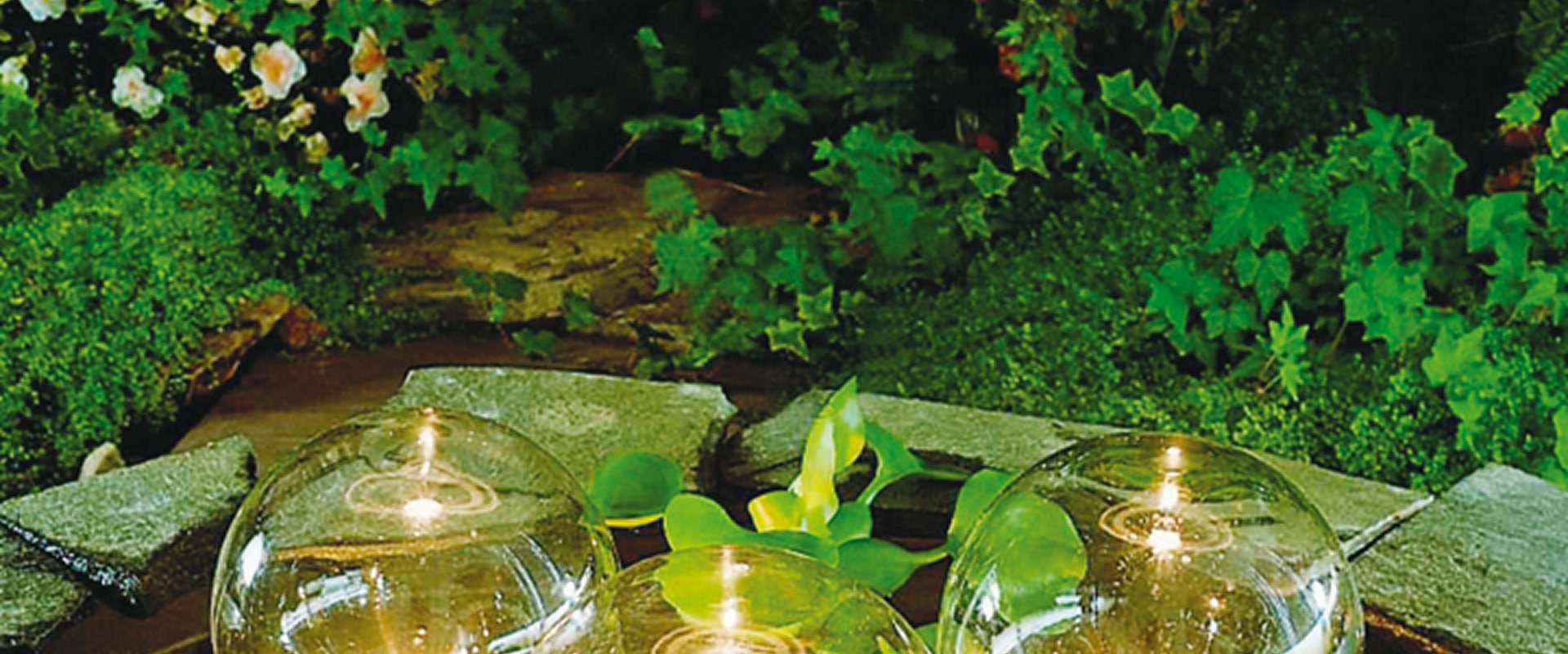 Jardin luxuriant avec mousse, lierre et fleurs pâles. Trois globes lumineux en verre posés sur des pierres, créant une ambiance paisible.