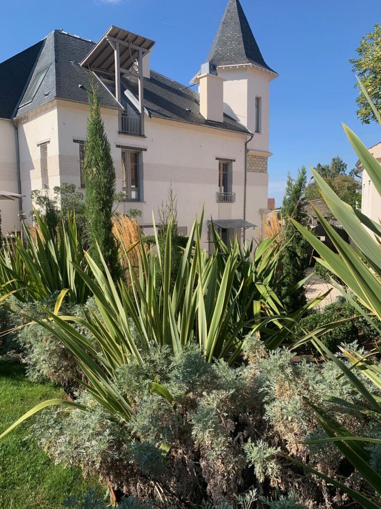 Bâtiment moderne avec tour et toit gris, entouré d'un jardin luxuriant de plantes vertes et argentées sous un ciel bleu clair.