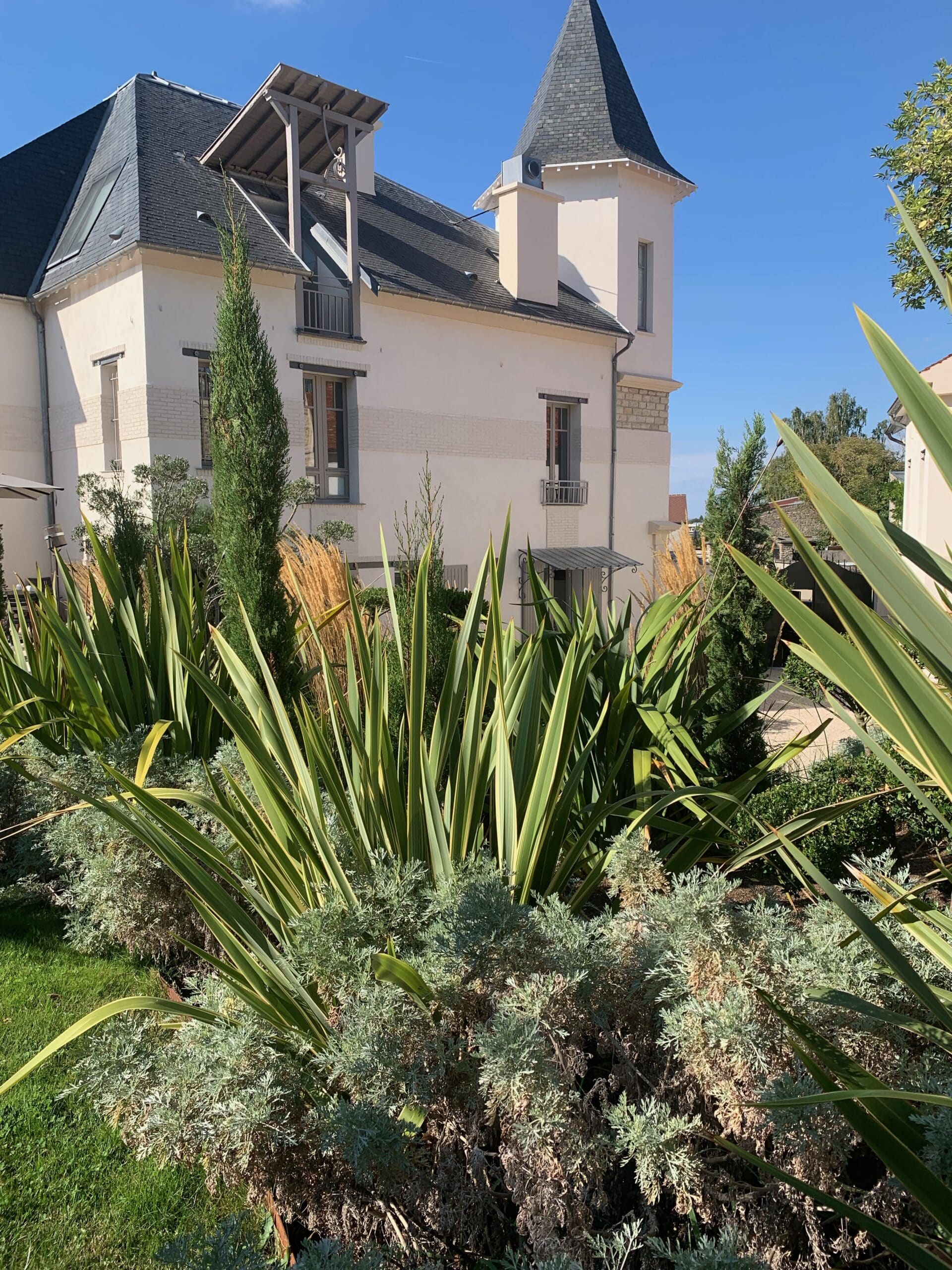 Bâtiment moderne avec tour et toit gris, entouré d'un jardin luxuriant de plantes vertes et argentées sous un ciel bleu clair.