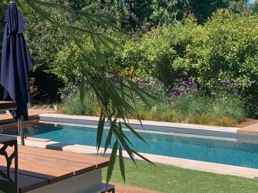 Grande piscine rectangulaire entourée de verdure luxuriante et d'une terrasse en bois. Un parasol sombre est replié près de l'eau.