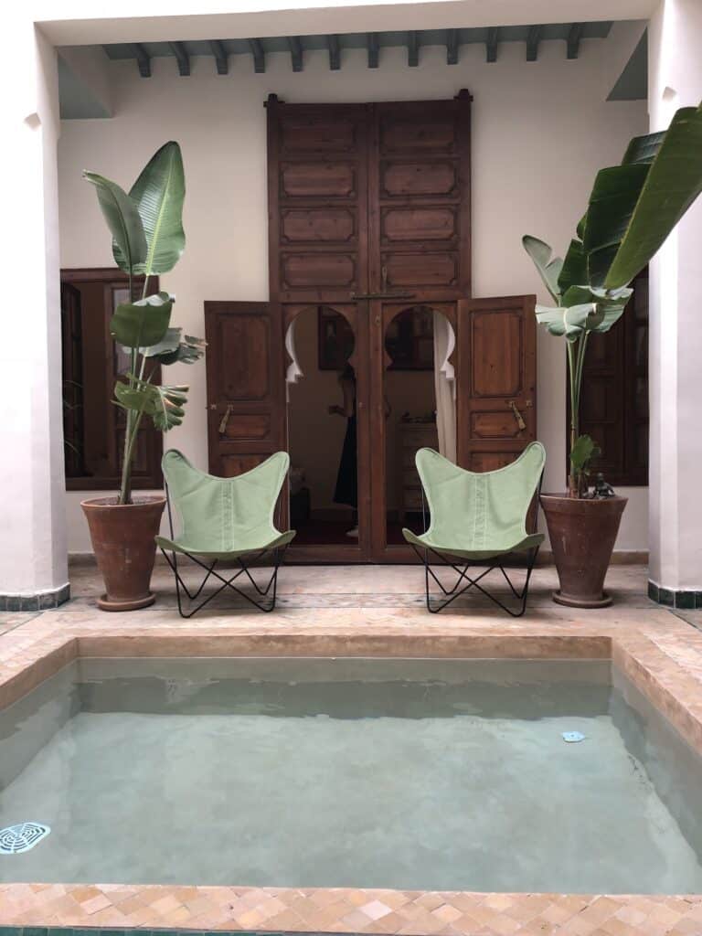 Riad marocain avec bassin central. Deux chaises papillon vertes, grandes plantes et portes en bois ouvertes. Personne visible à l'intérieur.