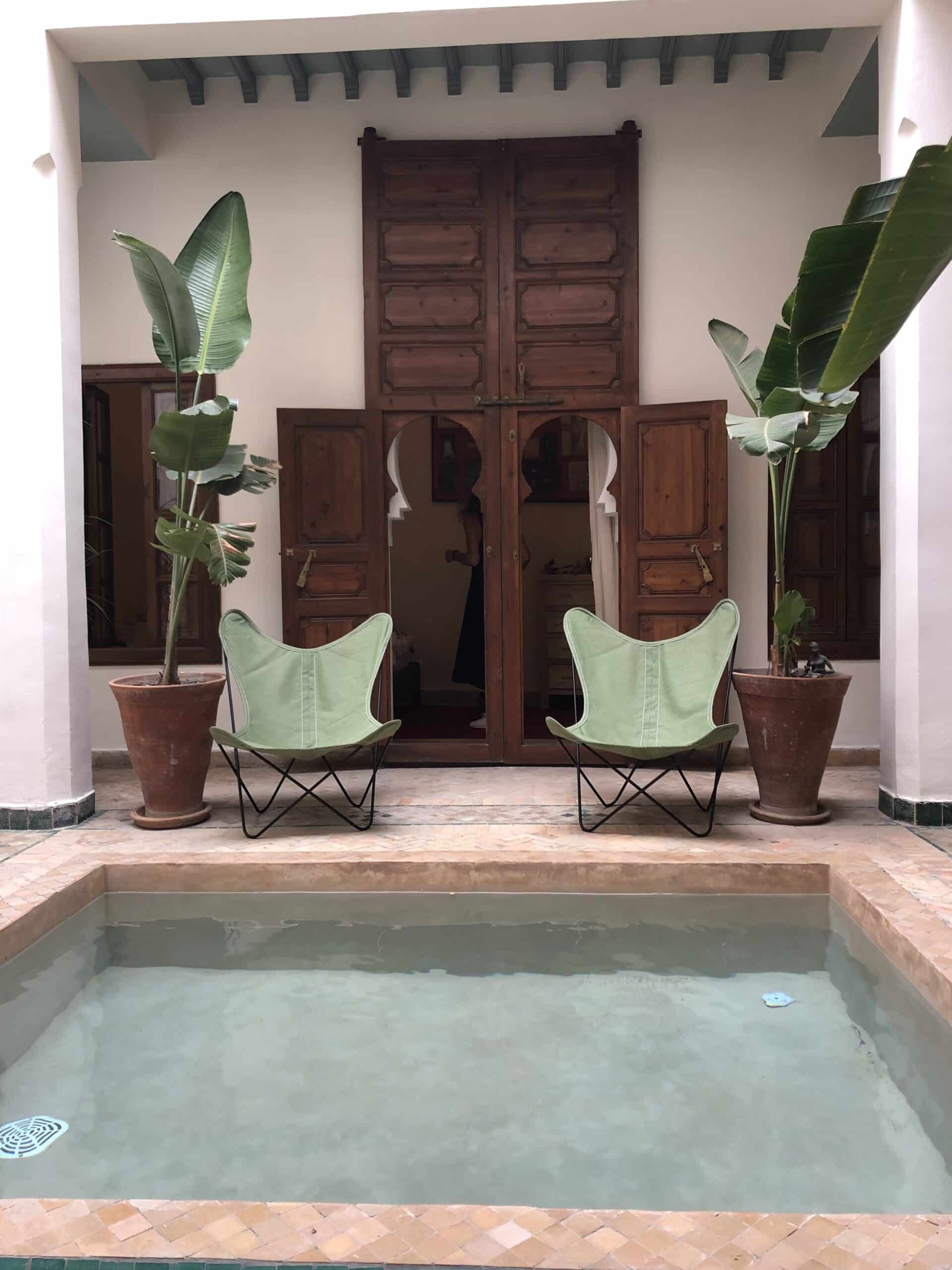 Riad marocain avec bassin central. Deux chaises papillon vertes, grandes plantes et portes en bois ouvertes. Personne visible à l'intérieur.