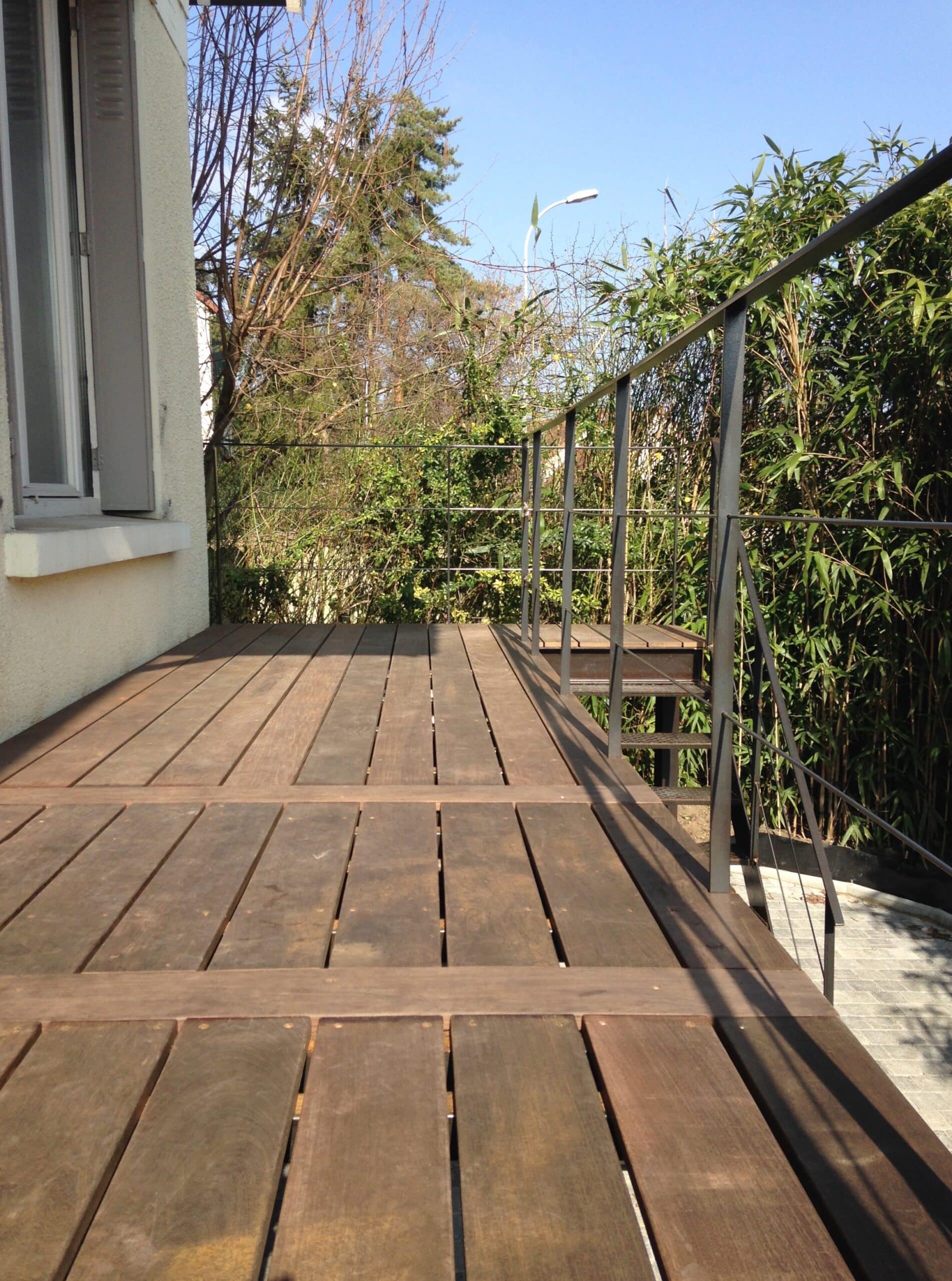 Terrasse en bois foncé avec balustrade métallique gris anthracite. Des bambous luxuriants et des arbres encadrent le jardin extérieur sous un ciel bleu clair.