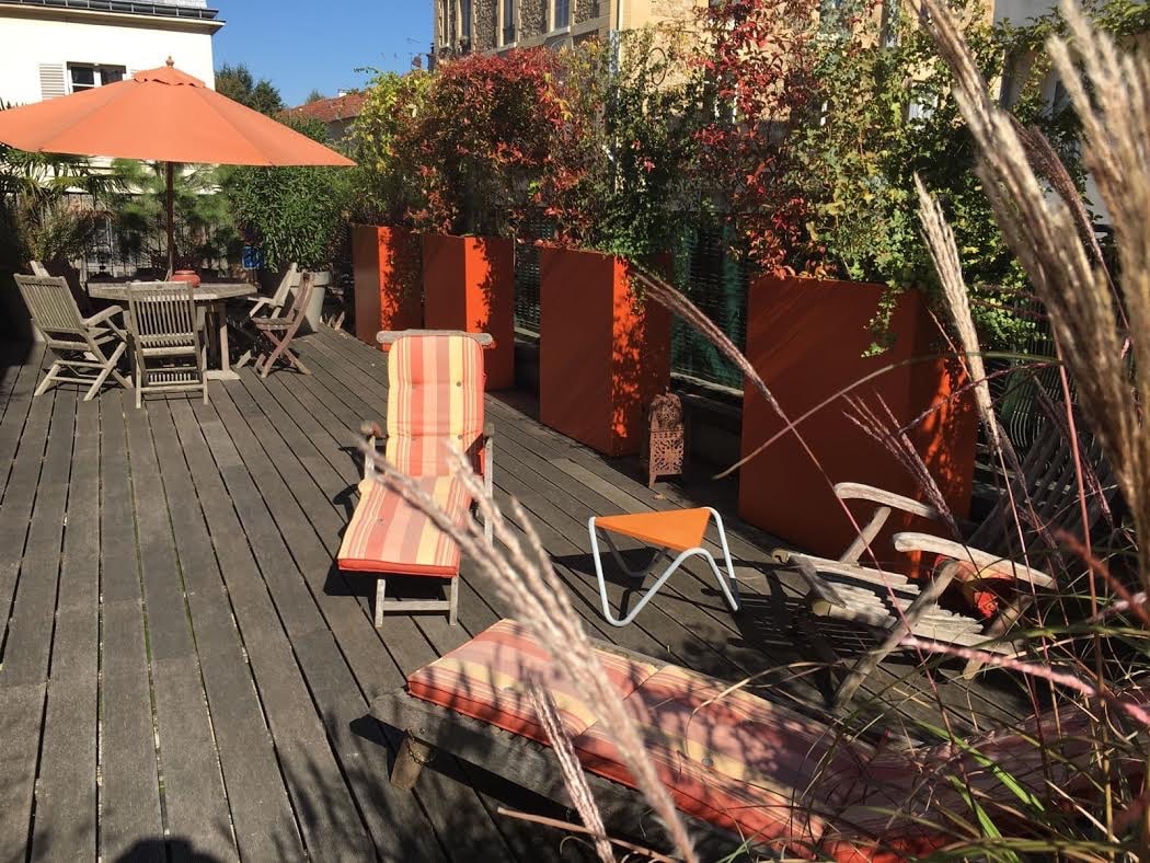 Terrasse en bois ensoleillée avec salon de jardin, parasol orange, transats rayés et grands bacs à plantes luxuriants.