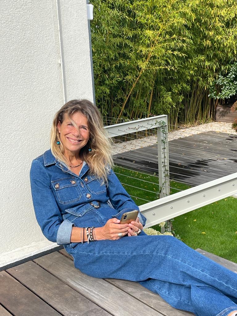 Femme souriante en combinaison jean sur terrasse en bois, tenant un smartphone. Arrière-plan de bambous verts.