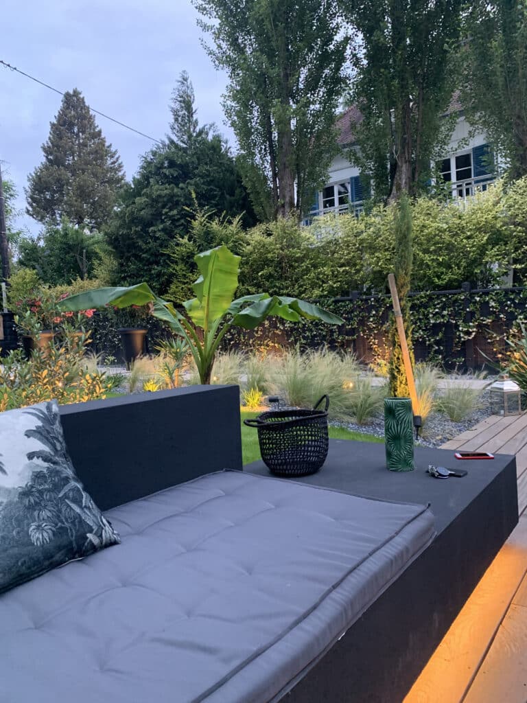 Salon de jardin moderne au crépuscule : canapé gris, éclairage chaud. Bananier, herbes ornementales et clôture fleurie en arrière-plan luxuriant.