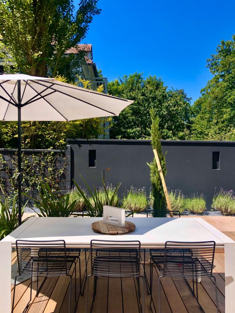 Terrasse moderne et ensoleillée avec table blanche, chaises noires et parasol. Mur végétal, plantes et ciel bleu clair.