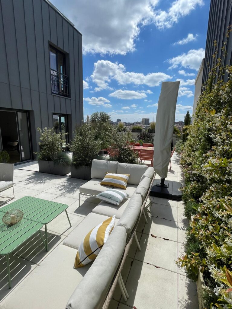 Terrasse de toit moderne avec canapé lounge, coussins rayés, table verte, plantes en pots et vue sur la ville sous ciel bleu.