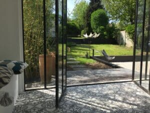 Luminosité d'un jardin luxuriant avec bambous, pelouse et terrasse, visible à travers des portes vitrées depuis une pièce aux carreaux ciment motifs gris.