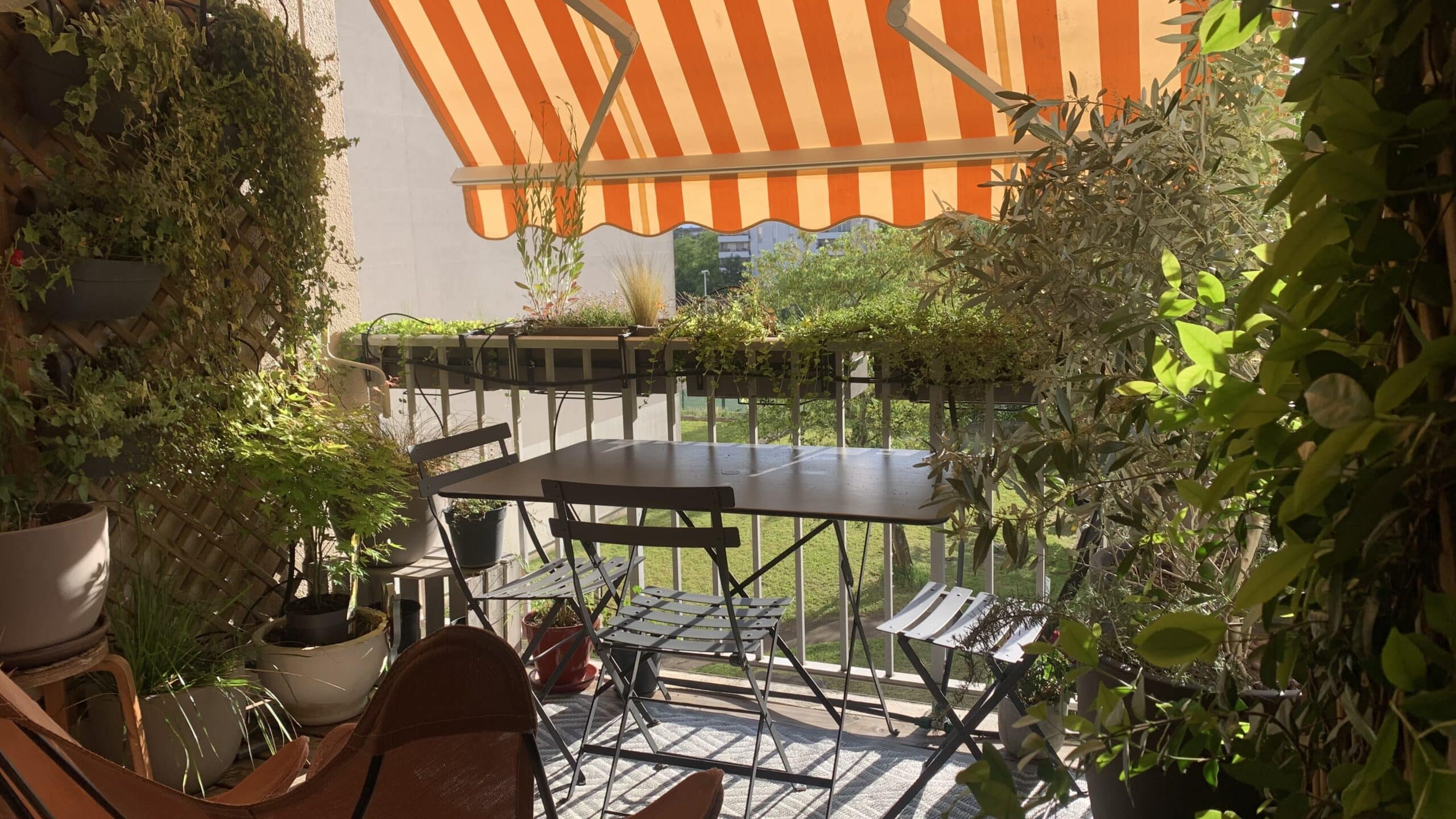 Balcon ensoleillé fleuri avec un auvent rayé orange et blanc, une table et des chaises noires, entouré de plantes vertes luxuriantes et de treillis.