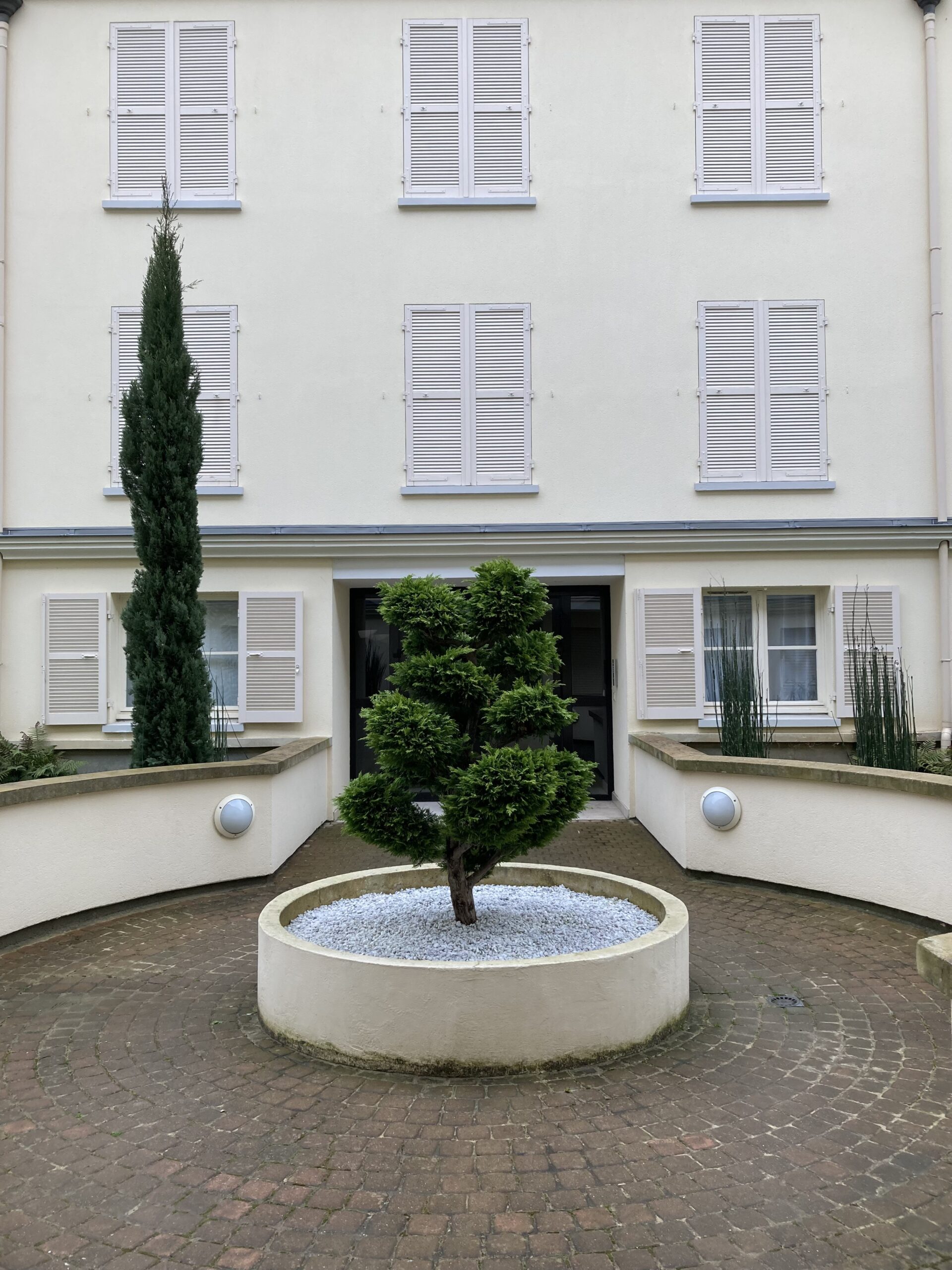 Façade crème à volets blancs, cour intérieure pavée de briques. Arbre topiaire central sur gravier blanc, grand cyprès, lumières murales.