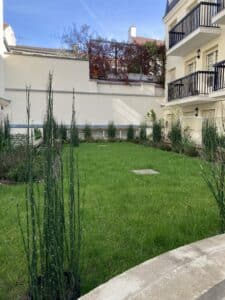 Cour intérieure résidentielle avec pelouse verte luxuriante, plantes en pots, murs crème, et balcons noirs. Ciel bleu lumineux.