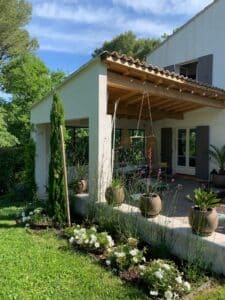 Maison méditerranéenne blanche, terrasse bois couverte. Jardin verdoyant avec rosiers blancs, cyprès, plantes en pots et pelouse sous ciel bleu.