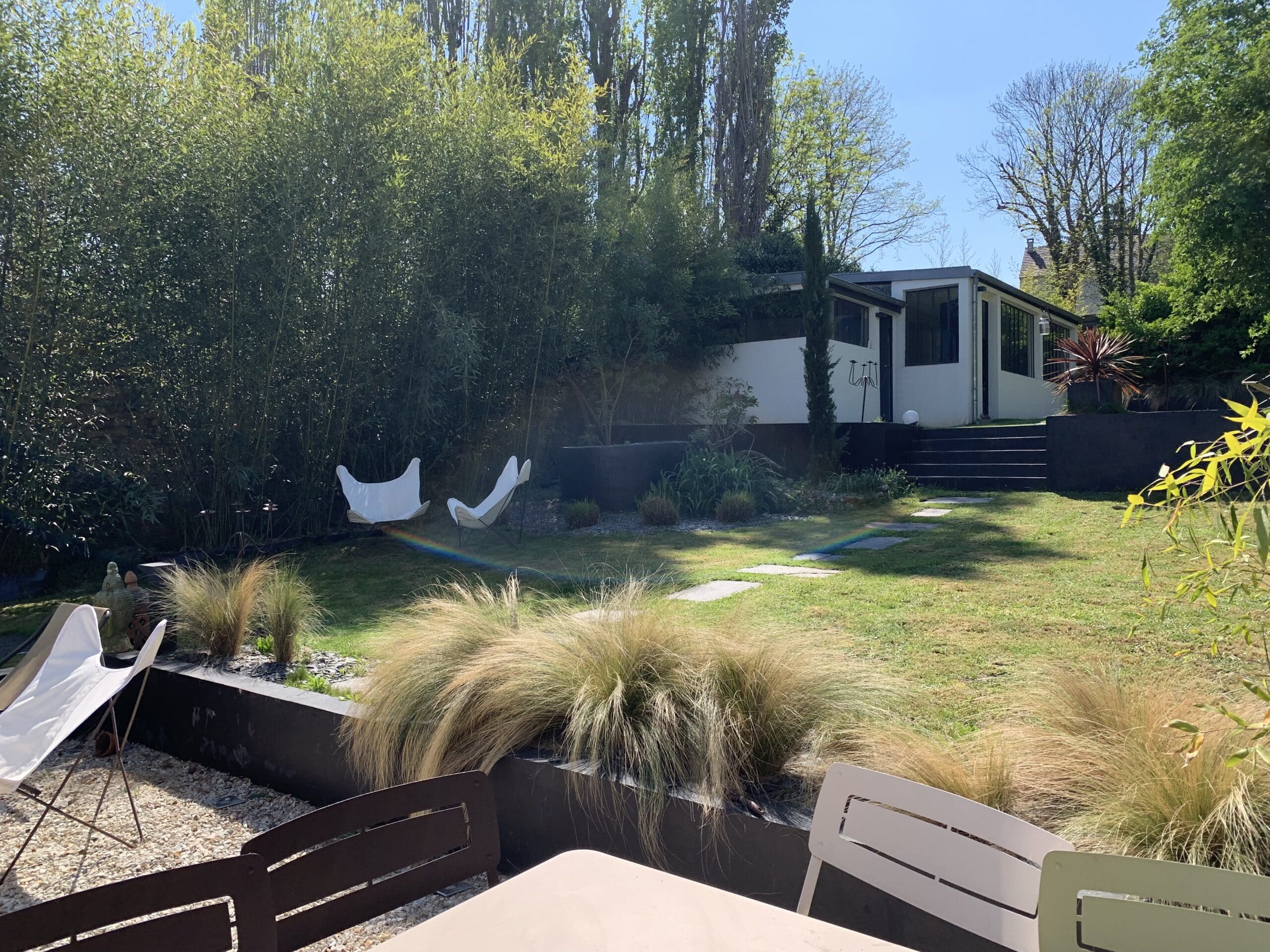 Jardin moderne ensoleillé: pelouse, chaises papillon blanches, herbes ornementales. Maison contemporaine à terrasses, bambous et arbres. Arc-en-ciel sur l'herbe.