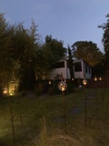 Jardin illuminé la nuit avec des lanternes suspendues et des orbes lumineuses sur des piquets, bordé de bambous luxuriants et d'une maison lointaine.