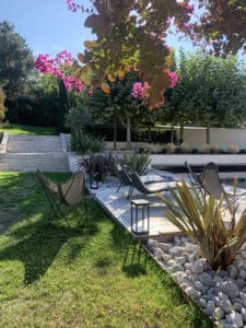 Jardin moderne avec piscine, terrasse en bois, chaises longues, plantes et fleurs roses éclatantes sous un ciel bleu ensoleillé.