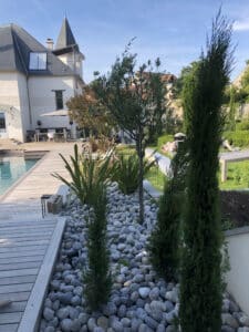 Piscine azur et terrasse en bois clair. Jardin paysagé zen avec galets blancs, olivier et plantes. Grande maison élégante sous ciel bleu.