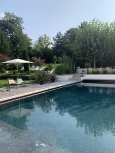 Piscine moderne bleu-vert avec terrasse en bois, deux chaises longues et un parasol blanc. Grand jardin luxuriant et ciel clair.