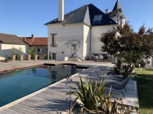 Grande maison moderne avec piscine à débordement et terrasse en bois. Mobilier de jardin, plantes vertes et ciel bleu clair.