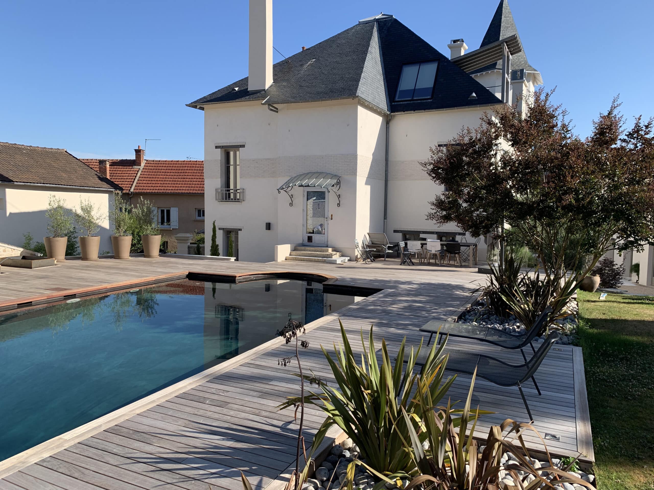 Grande maison moderne avec piscine à débordement et terrasse en bois. Mobilier de jardin, plantes vertes et ciel bleu clair.