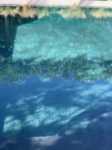 Eau turquoise d'une piscine ensoleillée, avec des reflets ondulants de ciel et de verdure. Le fond clair et des marches incurvées sont visibles.