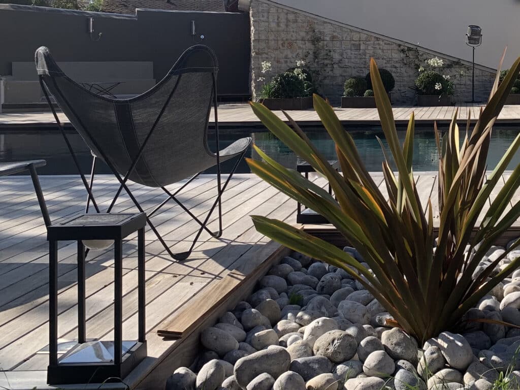 Terrasse moderne en bois avec chaise papillon noire, piscine, plante exotique et galets. Mur en pierre et maison contemporaine.