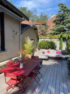 Terrasse en bois avec table rouge, chaises et canapé extérieur gris à coussins rouges. Verdure luxuriante et ciel bleu d'été.