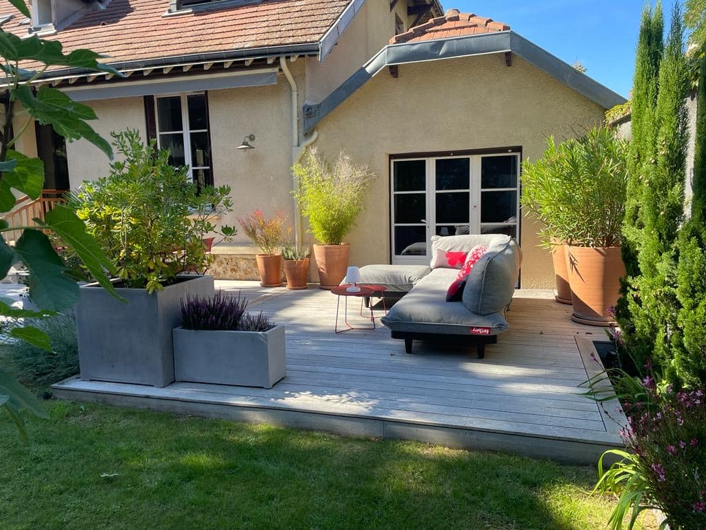 Terrasse en bois ensoleillée avec chaise longue Fatboy grise, coussins rouges, et diverses plantes en pots. Maison beige et jardin luxuriant sous un ciel bleu clair.