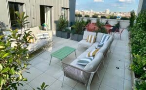 Terrasse de toit moderne avec canapés extérieurs beiges, coussins rayés jaunes et verts, table basse verte. Verdure luxuriante et vue dégagée sur la ville.