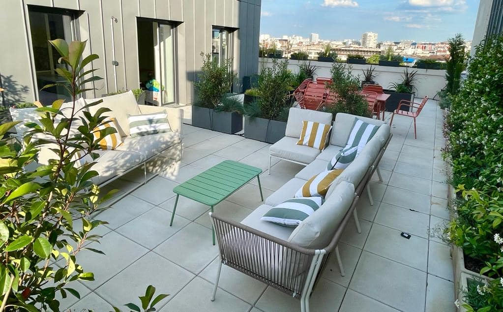Terrasse de toit élégante avec mobilier lounge beige, coussins rayés verts/jaunes, table basse verte. Plantes et vue ville ensoleillée.