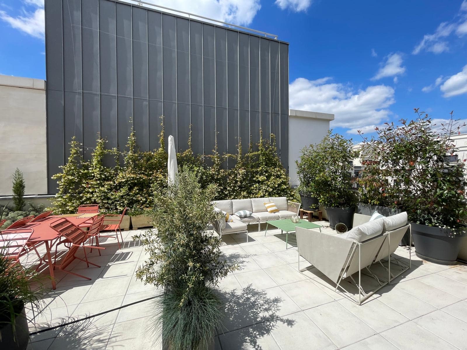 Spacieuse terrasse sur le toit avec mobilier de jardin moderne, sofa beige, table rouge, plantes en pots luxuriantes et ciel bleu.